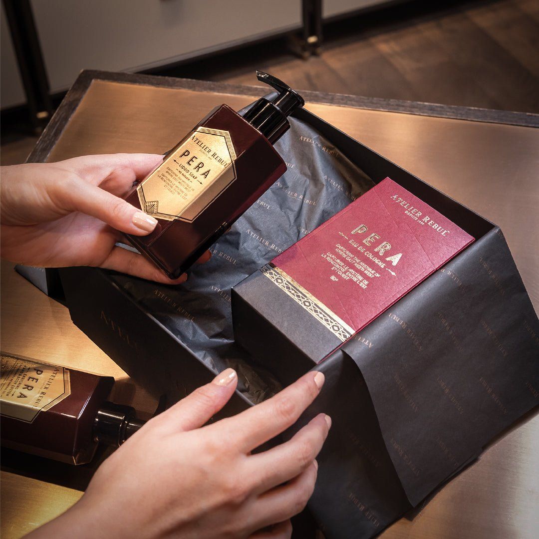 Hände halten eine braune Atelier Rebul Pera Handseife-Flasche und eine Schachtel. Die Schachtel enthält eine weitere Flasche und eine rote Schachtel.