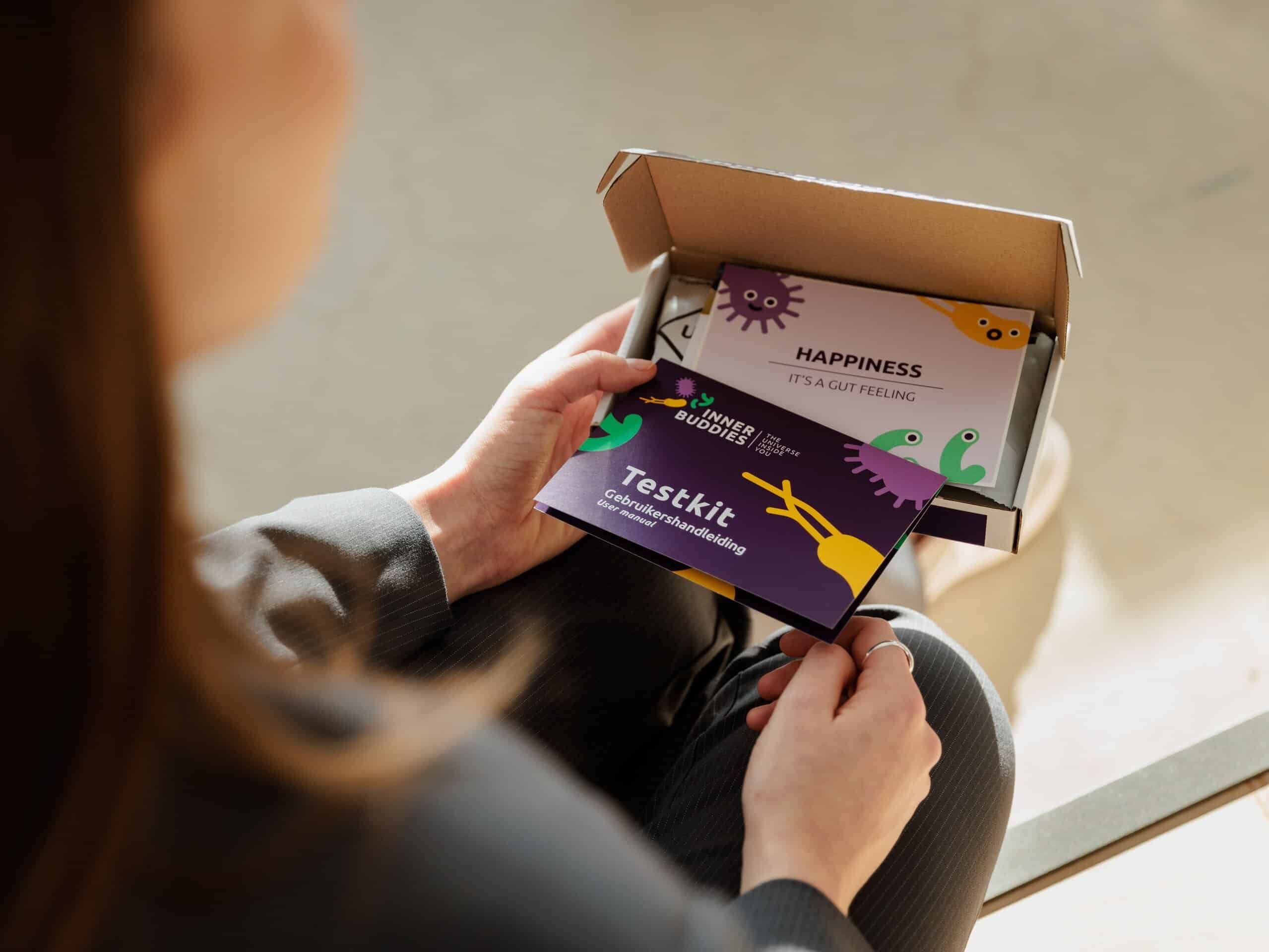 Person hält eine geöffnete Inner Buddies Box und eine Broschüre mit dem Titel „Testkit“ und Mikroben.