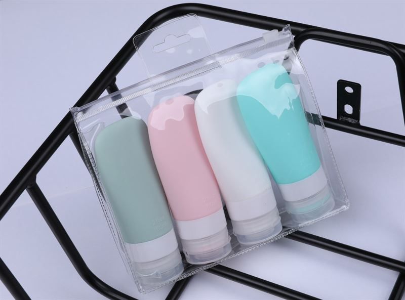 Vier farbige Reiseflaschen in einer transparenten Tasche. Farben: Grün, Rosa, Weiß und Türkis. Auf einem schwarzen Gestell.
