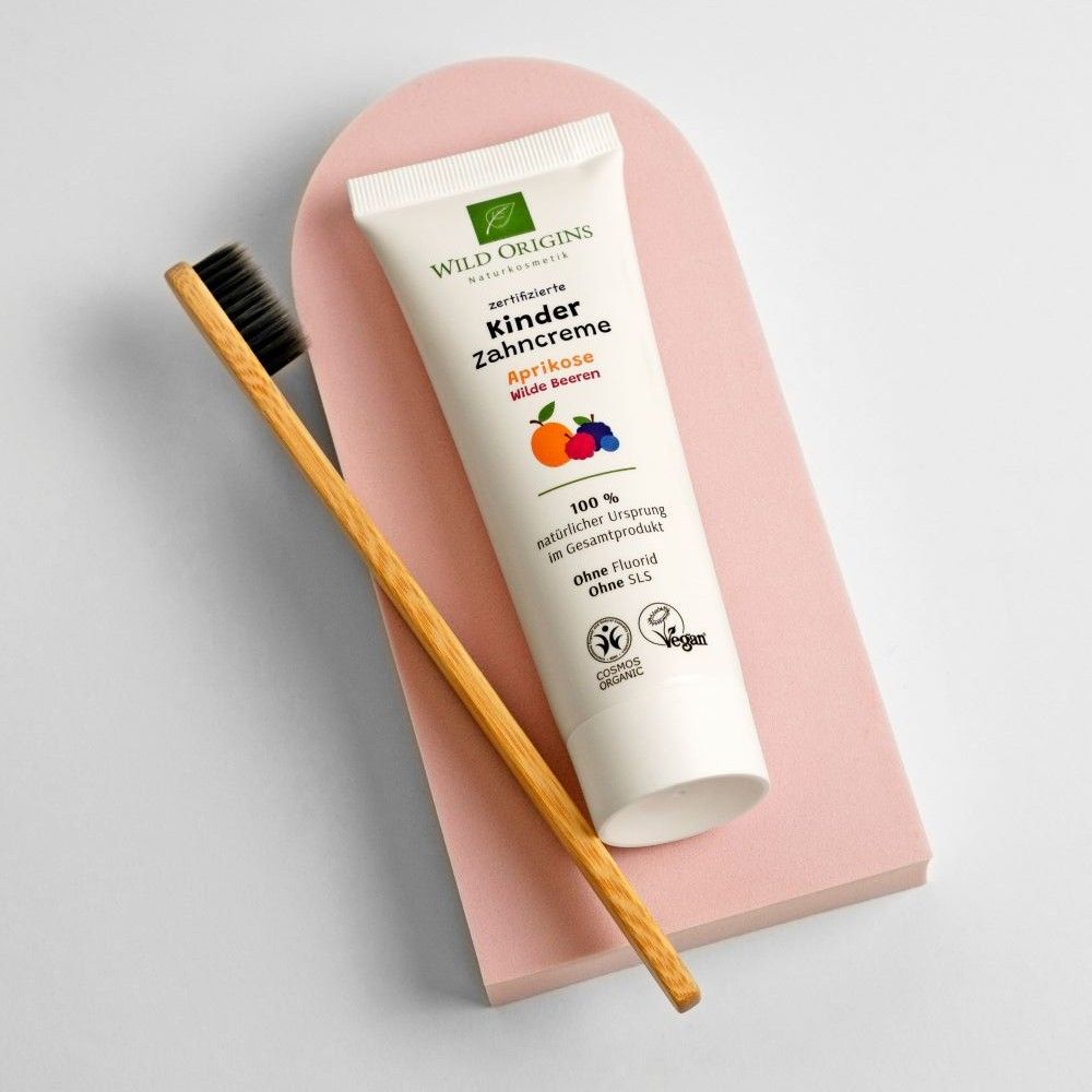 Zahncreme-Tube neben Holzzahnbürste auf rosa Podest. Produktbezeichnung und Logo sichtbar. Siegel für Naturkosmetik und Vegan.