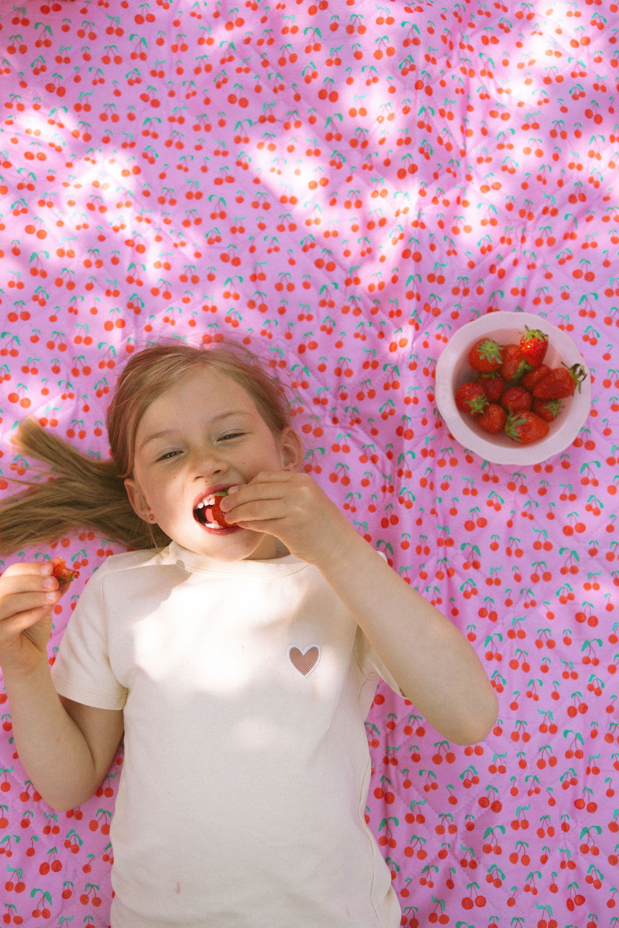 Kind liegt auf einer Decke mit Kirschmuster. Sie isst eine Erdbeere. Eine Schale mit Erdbeeren steht daneben.