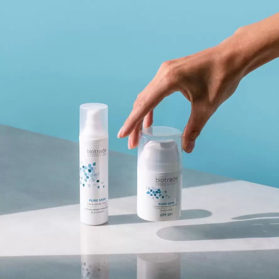 Zwei weiße Creme-Flaschen. Eine steht, die andere liegt. Aufschrift: Biotrade, Pure Skin, SPF 50+. Hand greift nach einer Flasche.