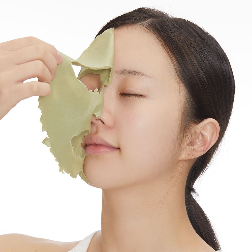 Gesicht einer Frau mit grüner Maske. Eine Hand zieht die Maske ab. Hintergrund weiß.