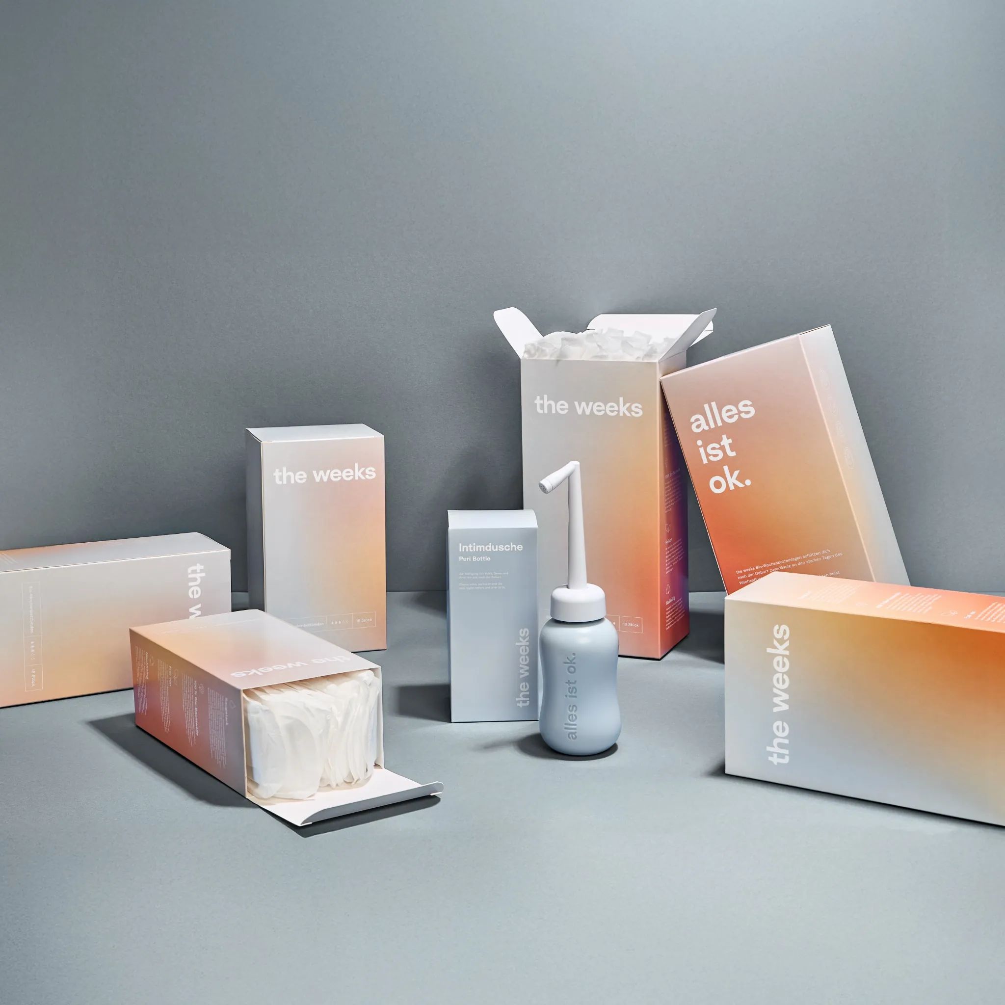 Mehrere Boxen mit dem Schriftzug "the weeks". Einige geöffnet, mit Inhalt. Farbverlauf von Weiß zu Orange.