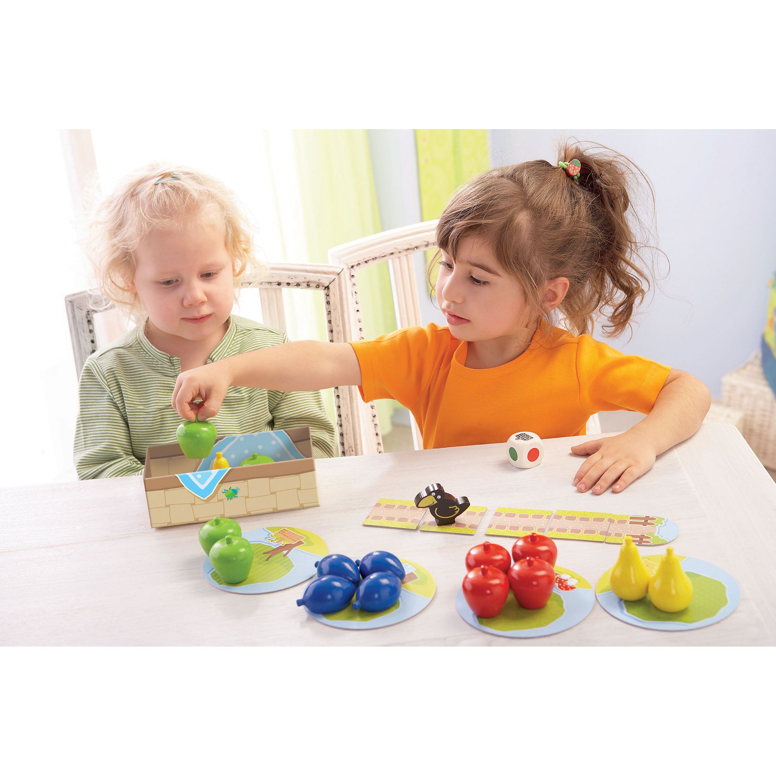 Zwei Kinder spielen das Spiel. Spielbrett, Obst, Würfel und Korb auf dem Tisch. Ein Kind legt Obst in den Korb.