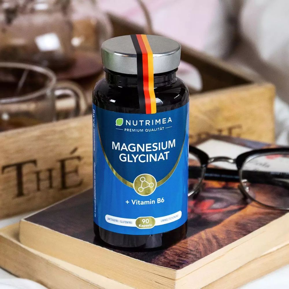 Flasche mit blauem Etikett auf Buch. Enthält Magnesium Glycinat + Vitamin B6. Mit deutschem Fähnchen.