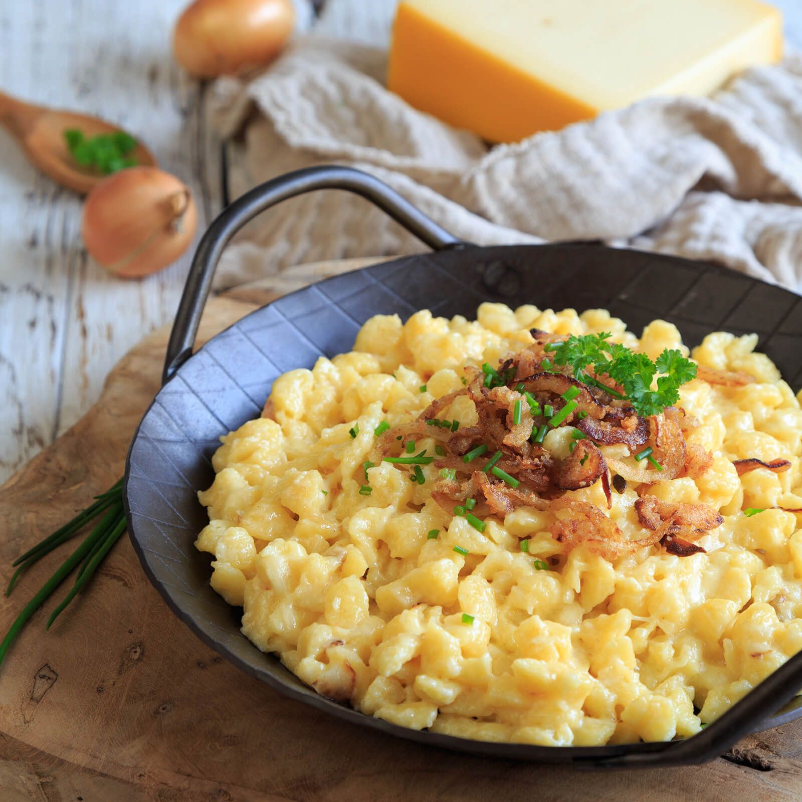 Pfanne mit Spätzle, garniert mit Röstzwiebeln und Petersilie. Auf einem Holztisch mit Zwiebeln und Käse.