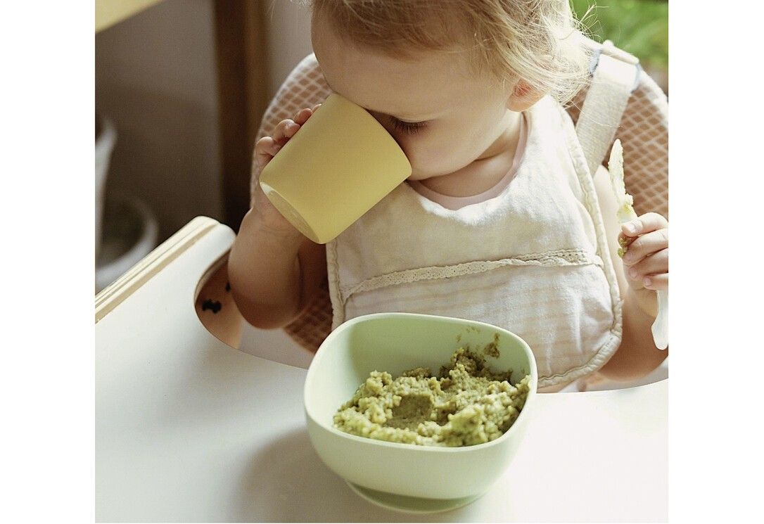 Kleinkind trinkt aus gelbem Becher. Grüne Schüssel mit Essen daneben. Geschirrset-Komponenten.