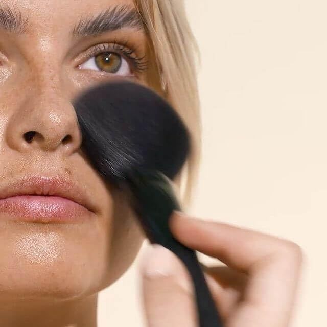 Person mit Pinsel, der Puder auf das Gesicht aufträgt. Heller Hintergrund.