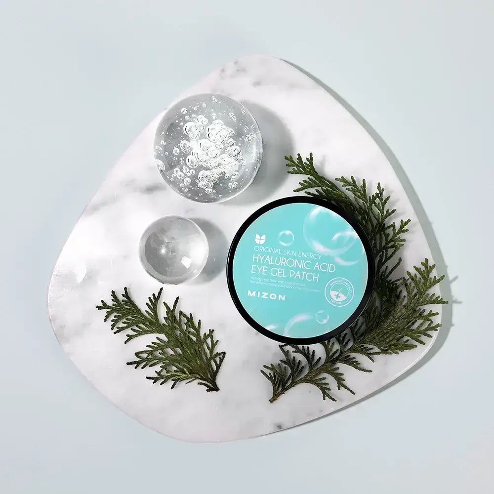 Runde Dose Augen Gel Patches mit blauem Deckel. Aufschrift: "Hyaluronsäure Eye Gel Patch". Dekoriert mit Zweigen und Glaskugeln.