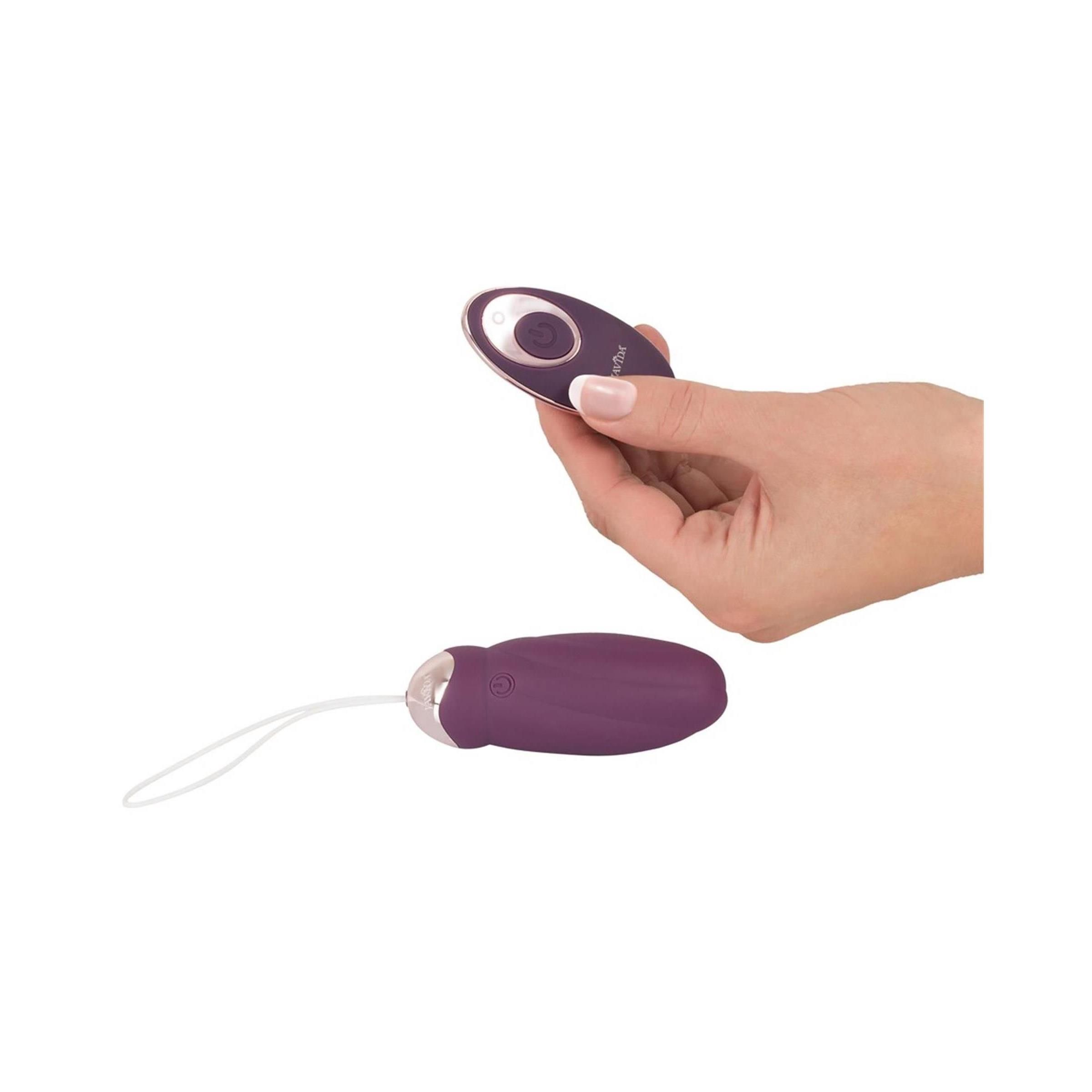 Hand hält Fernbedienung. Vibrationskugel und Fernbedienung liegen daneben. Beide in Lila und Gold.