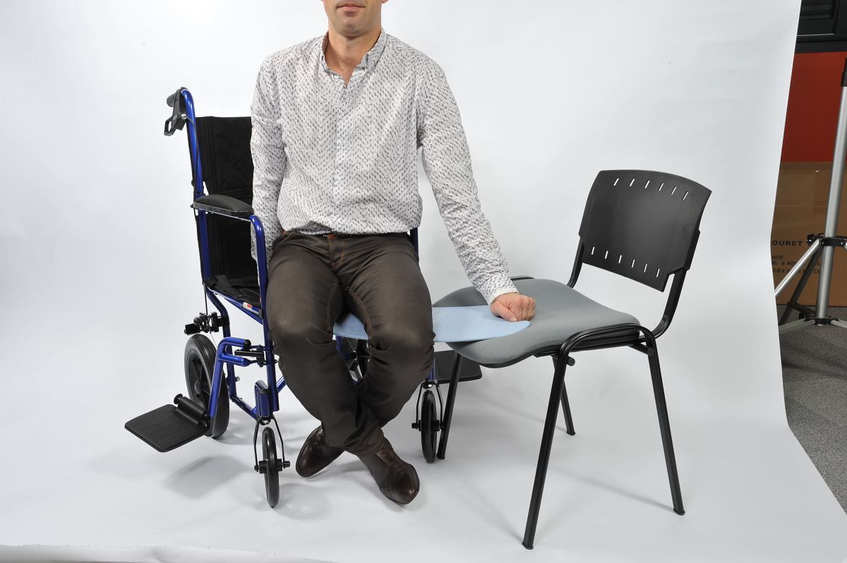 Transferbrett zwischen Rollstuhl und Stuhl. Person sitzt auf dem Brett. Rollstuhl links, Stuhl rechts.