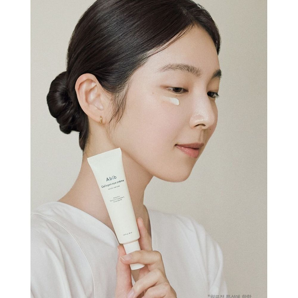 Frau hält eine hellbeige Creme-Tube. Aufschrift: Abib Collagen eye crème. Sie trägt die Creme auf die Haut unter dem Auge auf.
