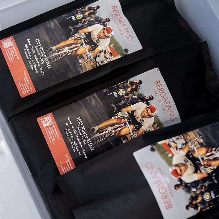 Drei schwarze Kaffeebohnen-Beutel mit Etikett. Aufdruck: BERGBRAND, Eddy Would Attack, Radfahrer-Illustration.