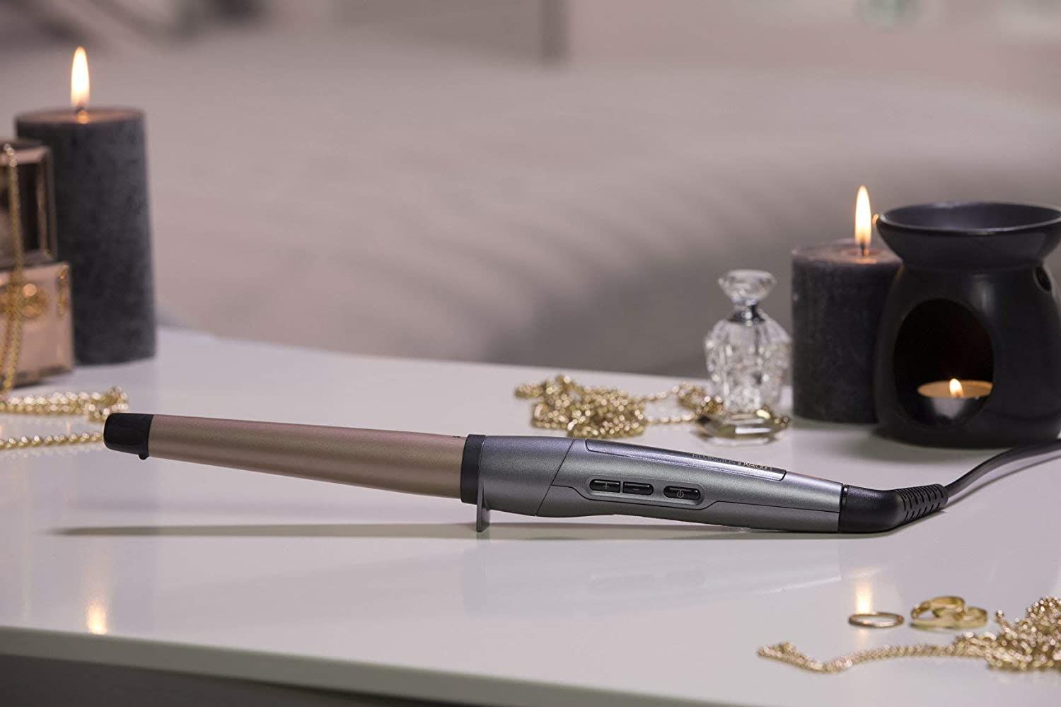 Lockenstab auf einem Tisch mit Kerzen, Schmuck und einem Duftdiffusor. Der Lockenstab hat einen silbernen Griff und eine roségoldfarbene Spitze.