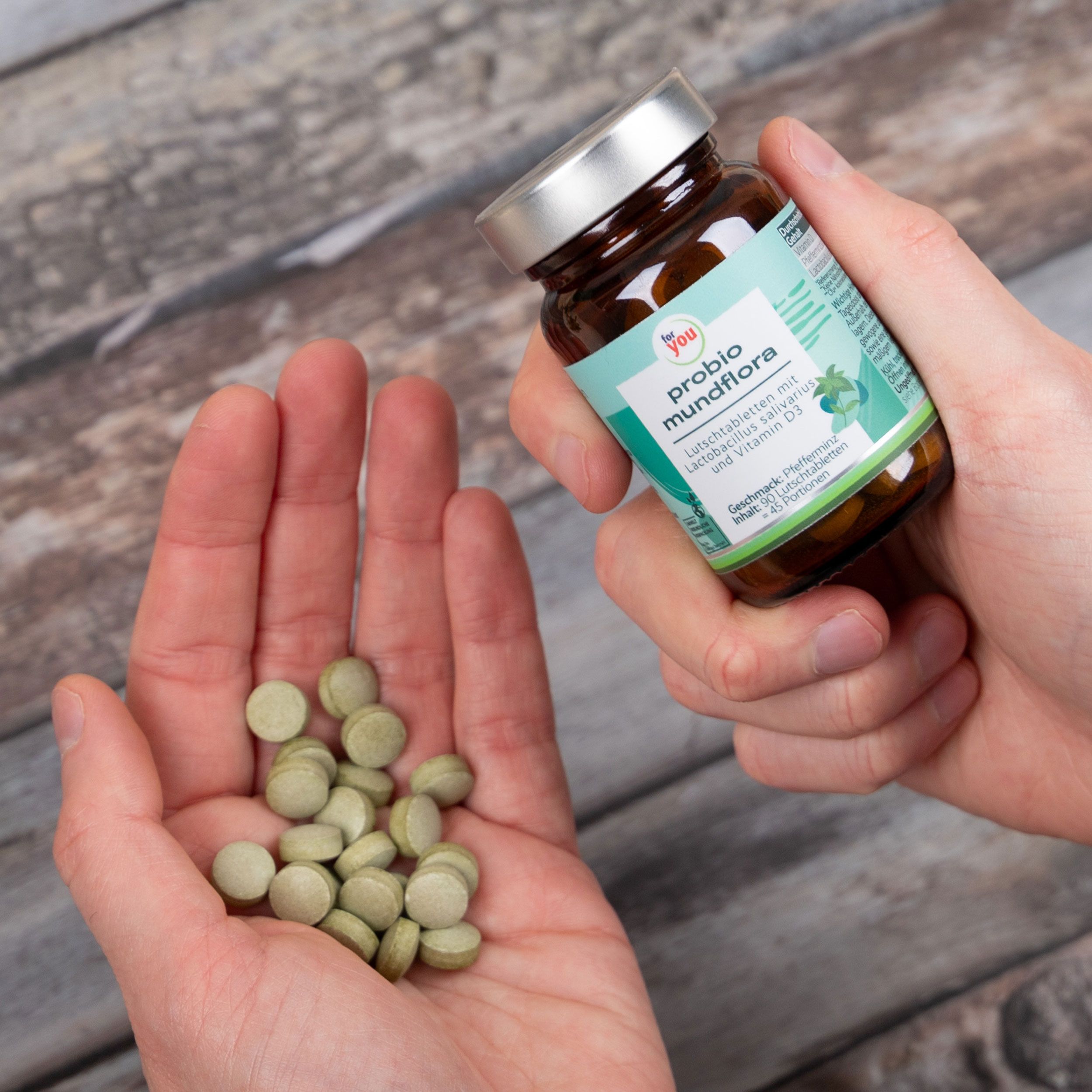 Hände halten Tabletten und eine Flasche. Tabletten grünlich. Aufschrift: probio mundflora. Braune Glasflasche.