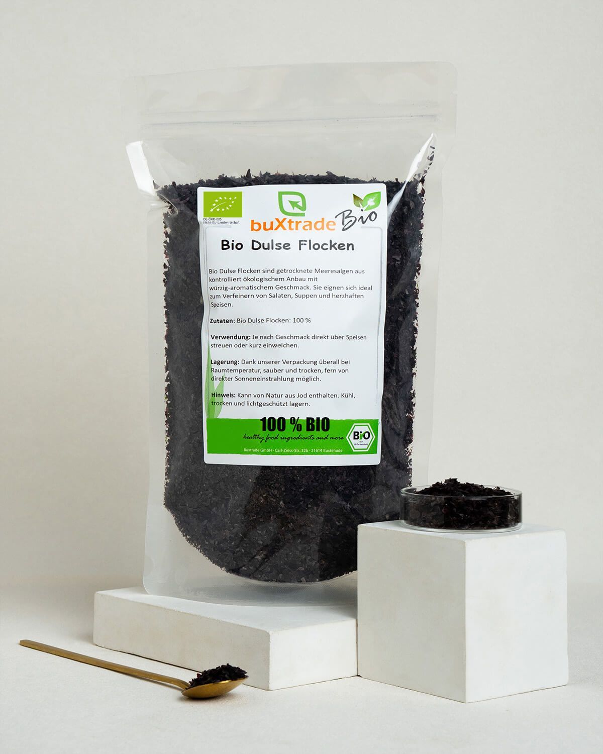 Ein Beutel Bio Dulse Flocken neben einem kleinen Behälter und einem Löffel mit Flocken. Weiße Podeste.