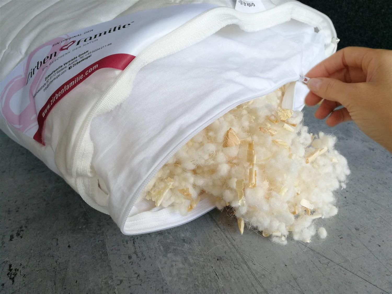 Weißes Kissen geöffnet, Füllung aus Holzspänen sichtbar. Hand öffnet Reißverschluss. Auf grauem Untergrund.