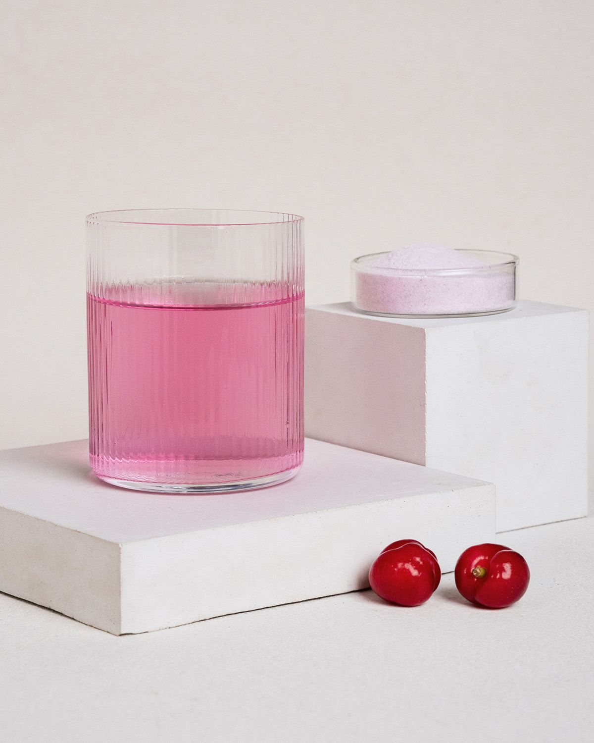 Glas mit rosa Flüssigkeit, Schale mit Pulver, zwei rote Früchte auf weißem Podest. Hintergrund hell.