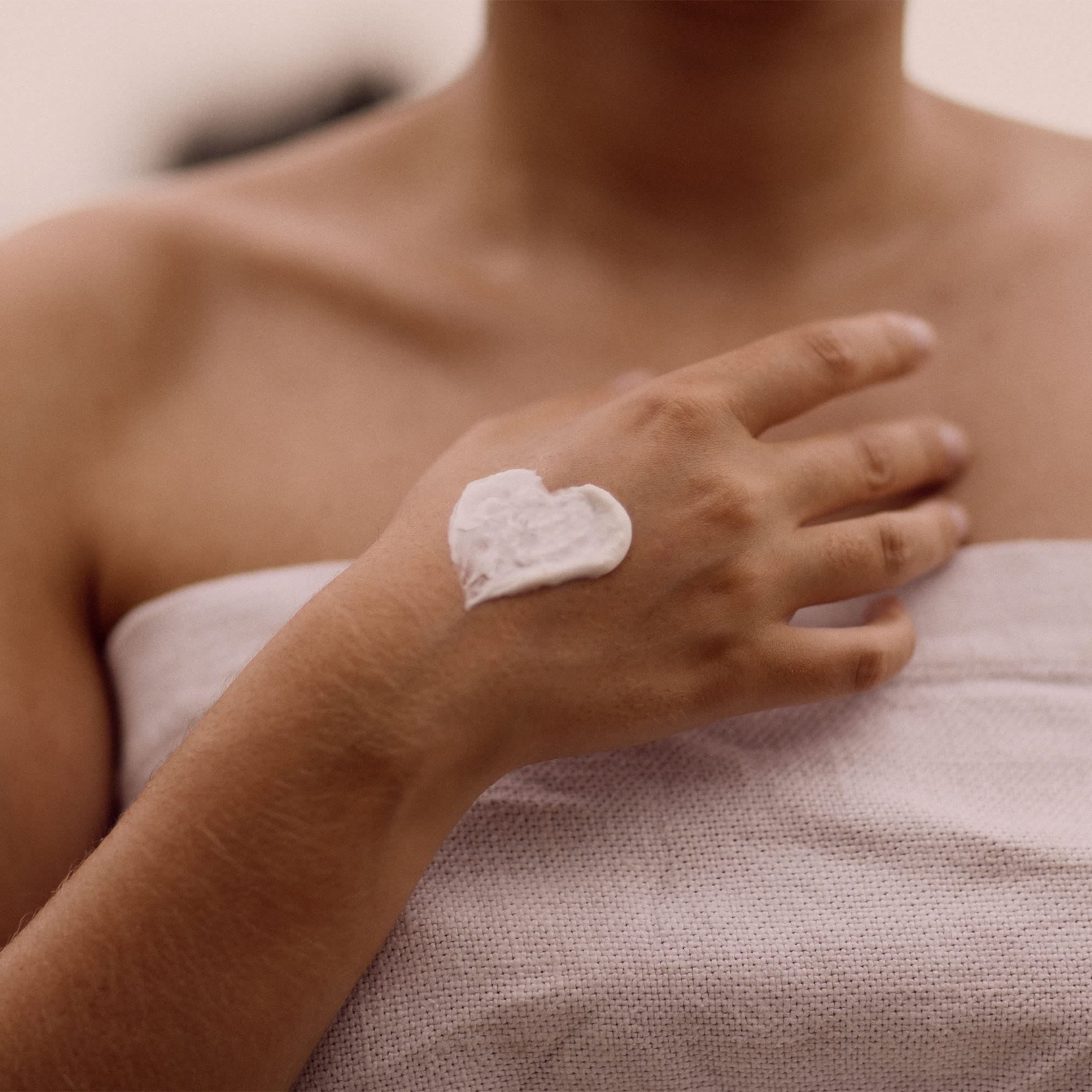 Hand mit Creme in Herzform. Person mit Handtuch bedeckt. Creme weiß.