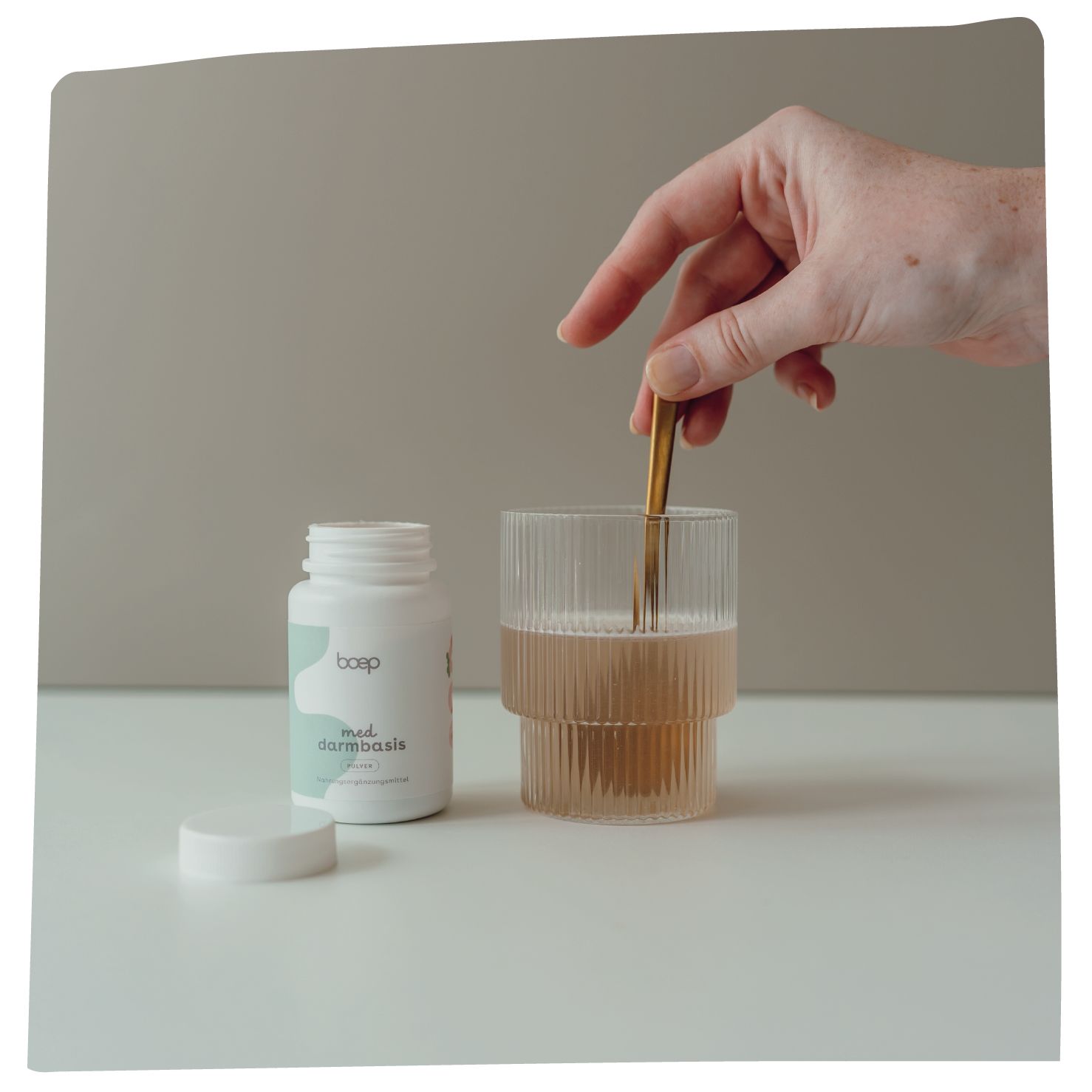 Weißes boep Med Darmbasis Pulver-Behältnis, Glas mit Flüssigkeit, Hand mit Löffel. Aufschrift und Logo sichtbar.