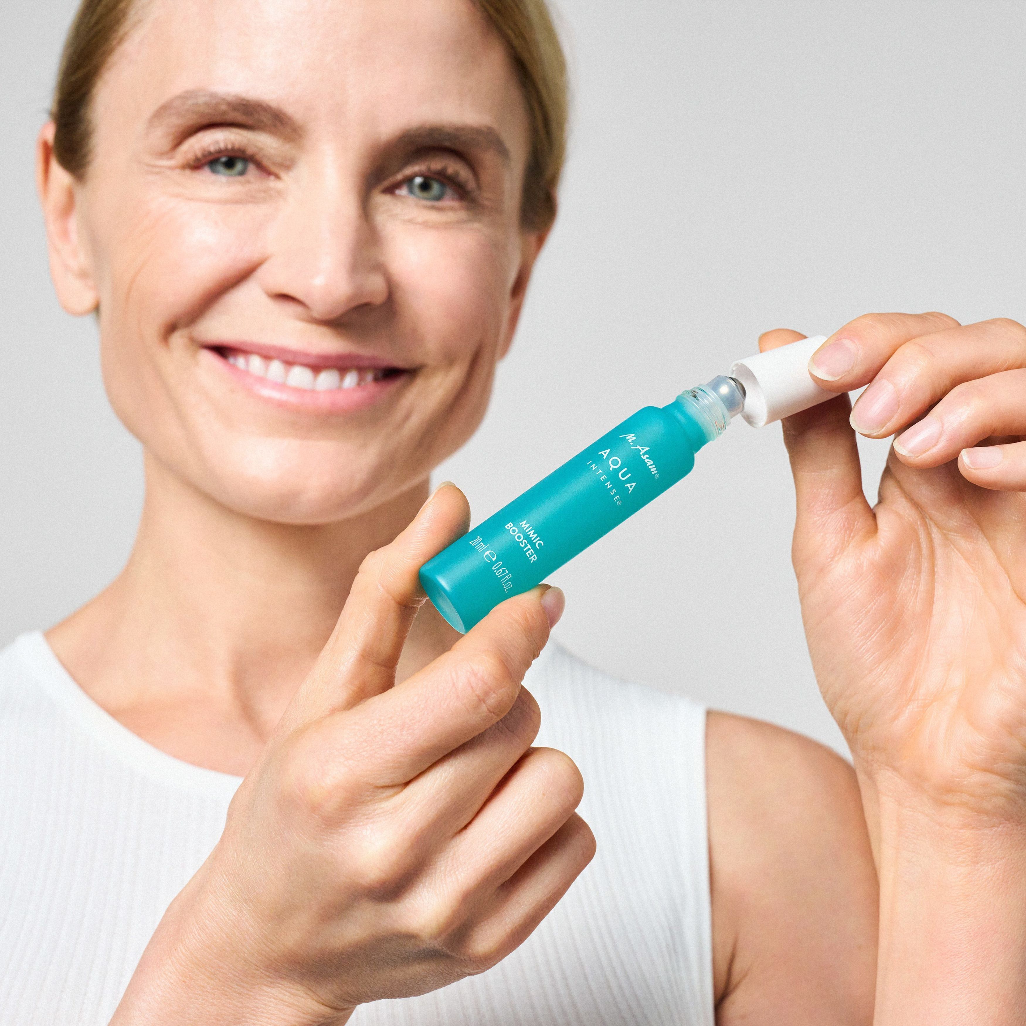 Frau hält eine Flasche. Aufschrift: M. Asam AQUA INTENSE Mimic Booster. Mit Roll-on-Applikator.
