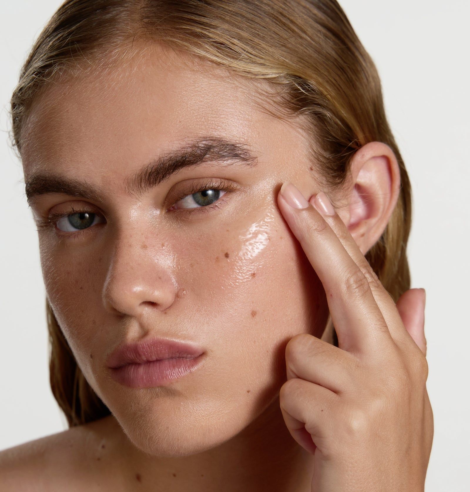 Gesicht einer Frau, die Serum auf die Haut aufträgt. Helle Haut, blonde Haare, Finger berühren die Wange. Serum sichtbar.