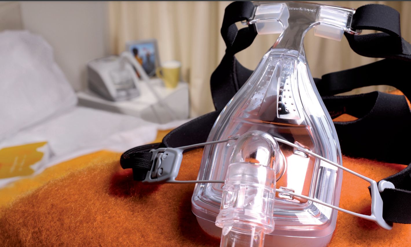 Atemmaske mit Kopfband auf einer Decke. Transparente Maske, schwarzes Kopfband. Im Hintergrund: Bett, Nachttisch.