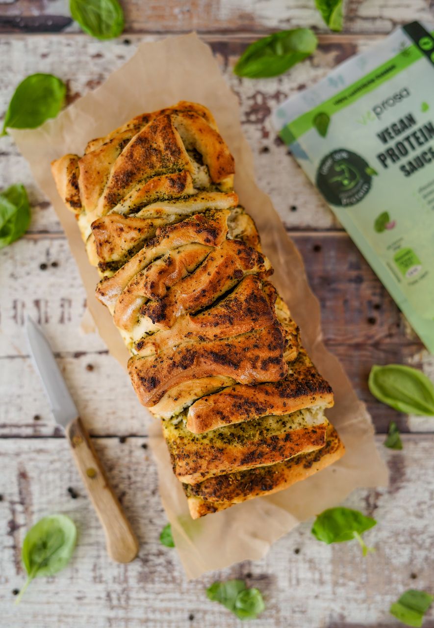 Geflochtener Brotlaib mit Pesto. Auf Backpapier, daneben Messer und Basilikumblätter. Im Hintergrund Verpackung von VEPROSA.