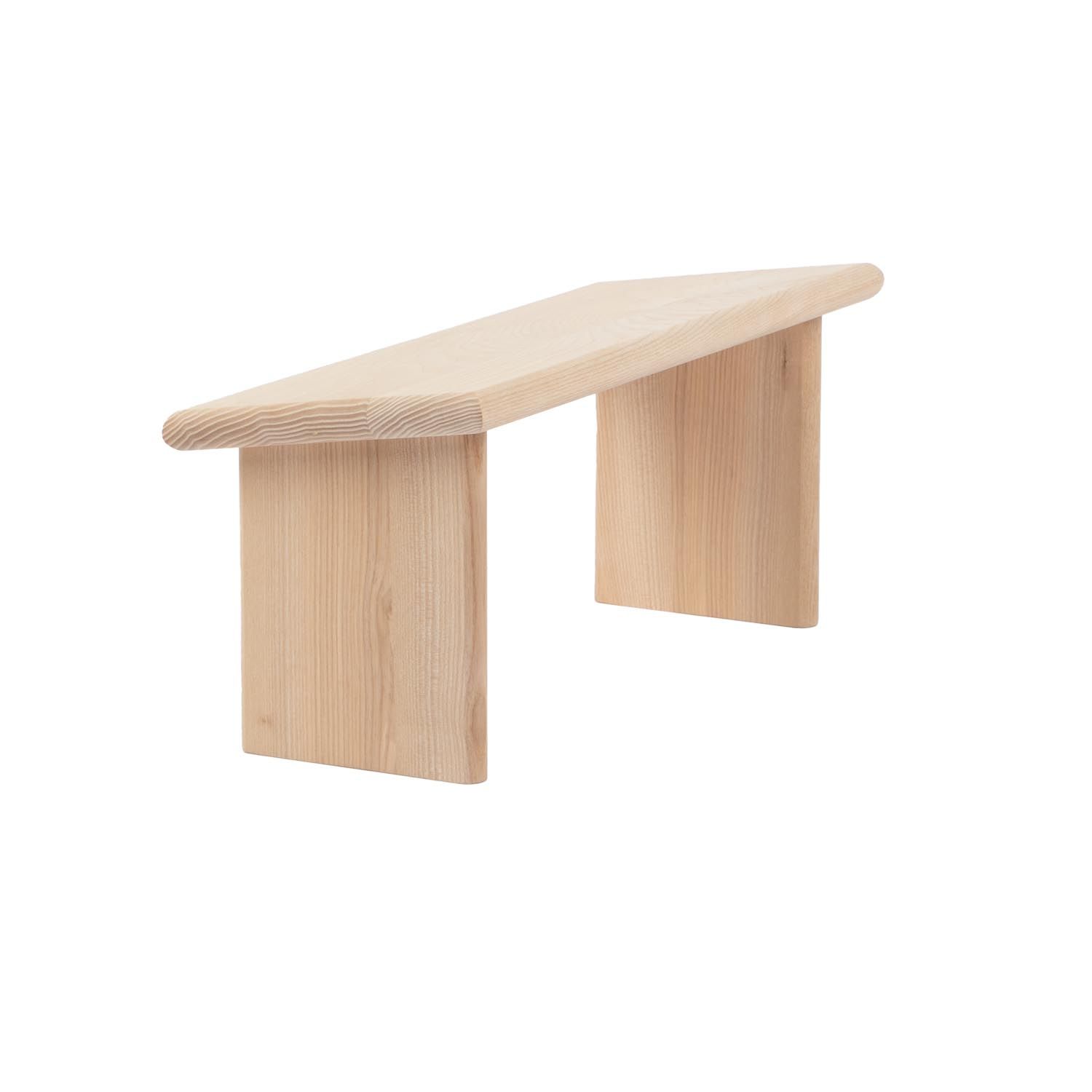 Hocker aus hellem Holz mit leicht geneigter Sitzfläche und zwei Beinen. Abgerundete Kanten.