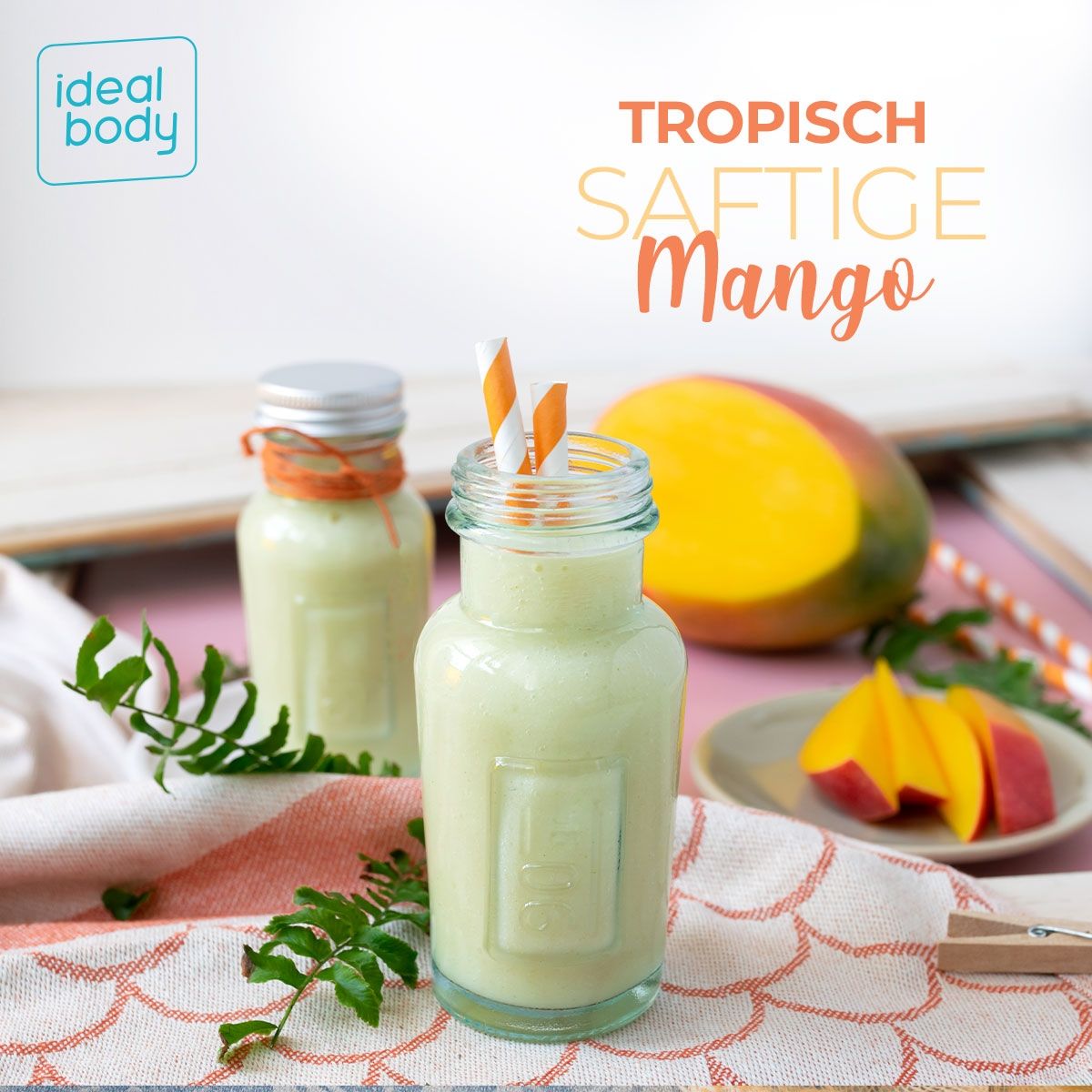 Zwei Glasflaschen mit Getränk, Strohhalme. Mango-Hälfte im Hintergrund. Text: "TROPISCH SAFTIGE Mango" und "ideal body"-Logo.