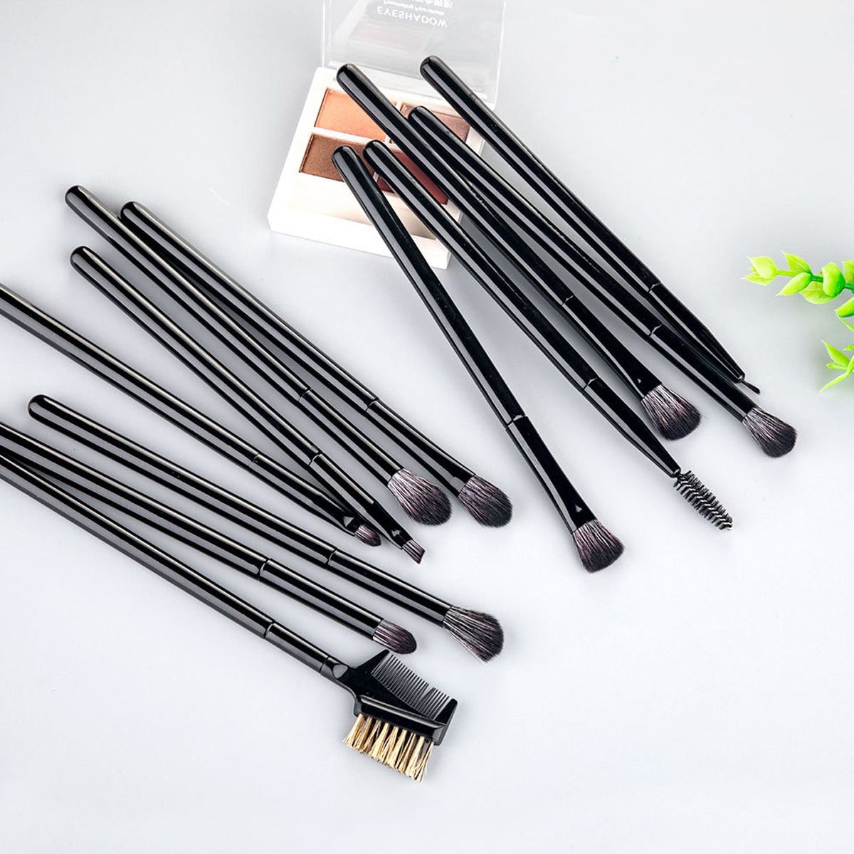 Schwarze Lidschatten- und Augenbrauenpinsel, teilweise auf einer grauen Fläche, mit einem Lidschatten-Set im Hintergrund.