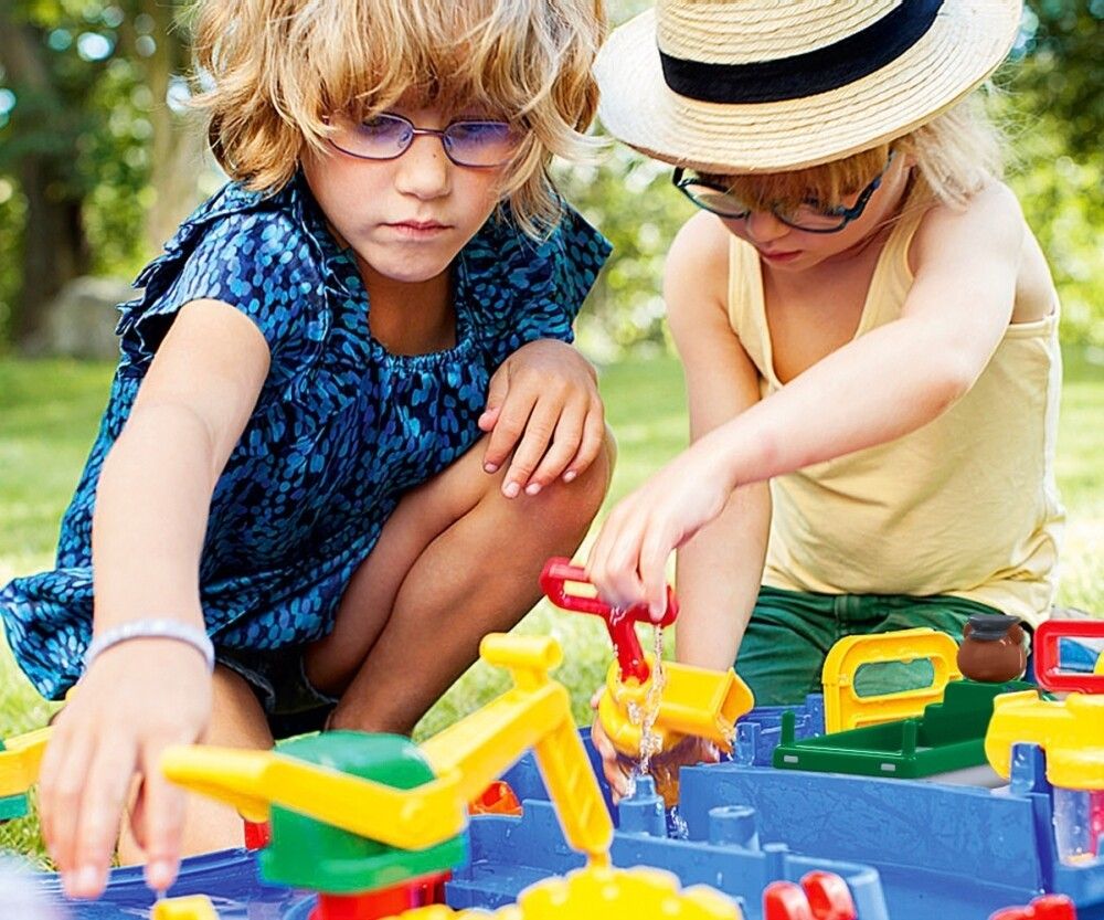 Zwei Kinder spielen mit einem AquaPlay-Set. Sie sitzen im Gras und interagieren mit dem Spielzeug. Wasser fließt.