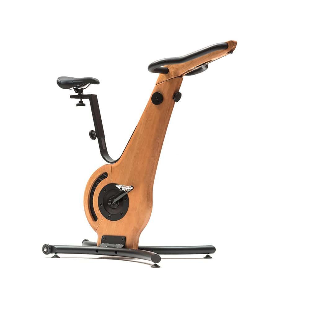 Ergometer aus Holz. Rahmen, Lenker und Sattel schwarz. Holzfarbener Hauptrahmen. Schwarze Pedale und Halterung. Auf schwarzem Sockel.