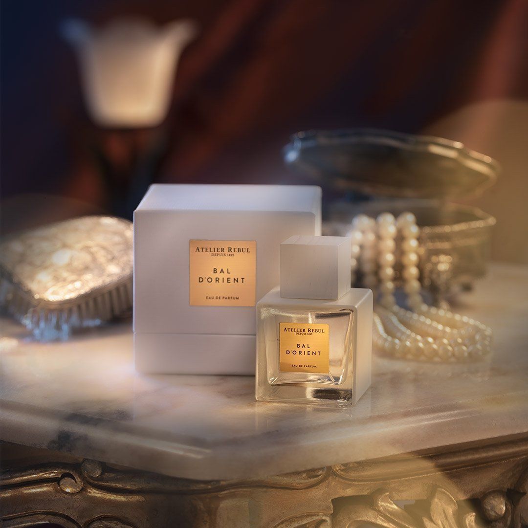 Parfümflakon und Verpackung. Eckiger Flakon mit hellem Inhalt, weißer Deckel. Goldfarbenes Etikett mit Schriftzug. Weiße Verpackung.