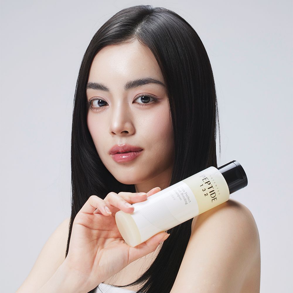Frau mit dunklem Haar hält eine Flasche COSRX Peptide 132 Ultra Perfect Hair Bonding Shampoo.