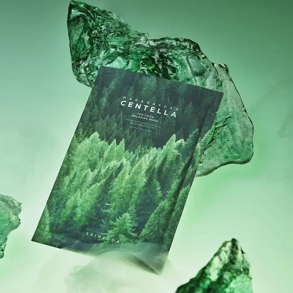 Verpackung der Gesichtsmaske mit Waldmotiv und Schriftzug MADAGASCAR CENTELLA. Liegt zwischen grünen Glasbrocken.