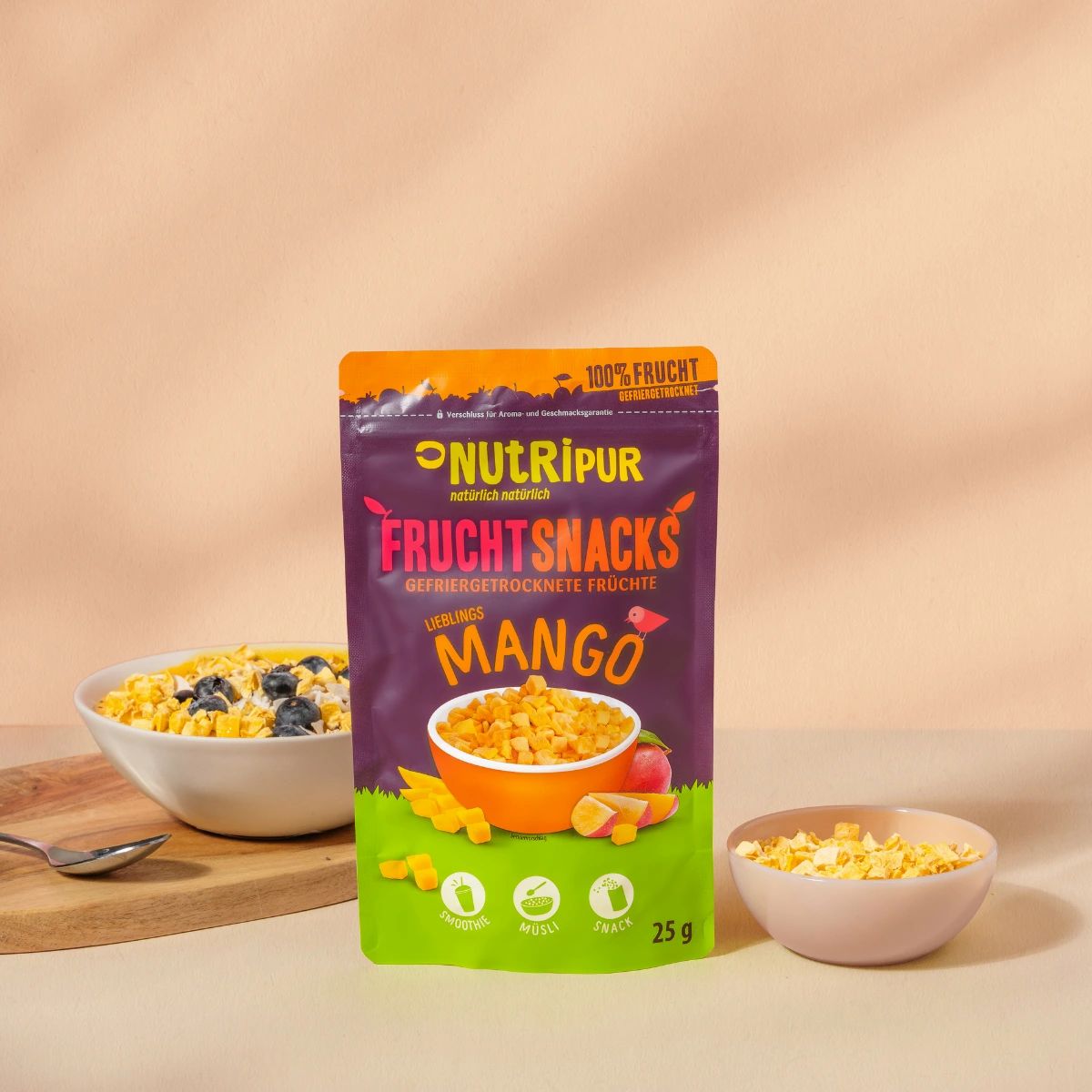 Verpackung von NutriPur Mango-Fruchtsnacks. Daneben Schalen mit Mango-Fruchtsnacks und Blaubeeren. Auf einem Holzbrett und einem Löffel.