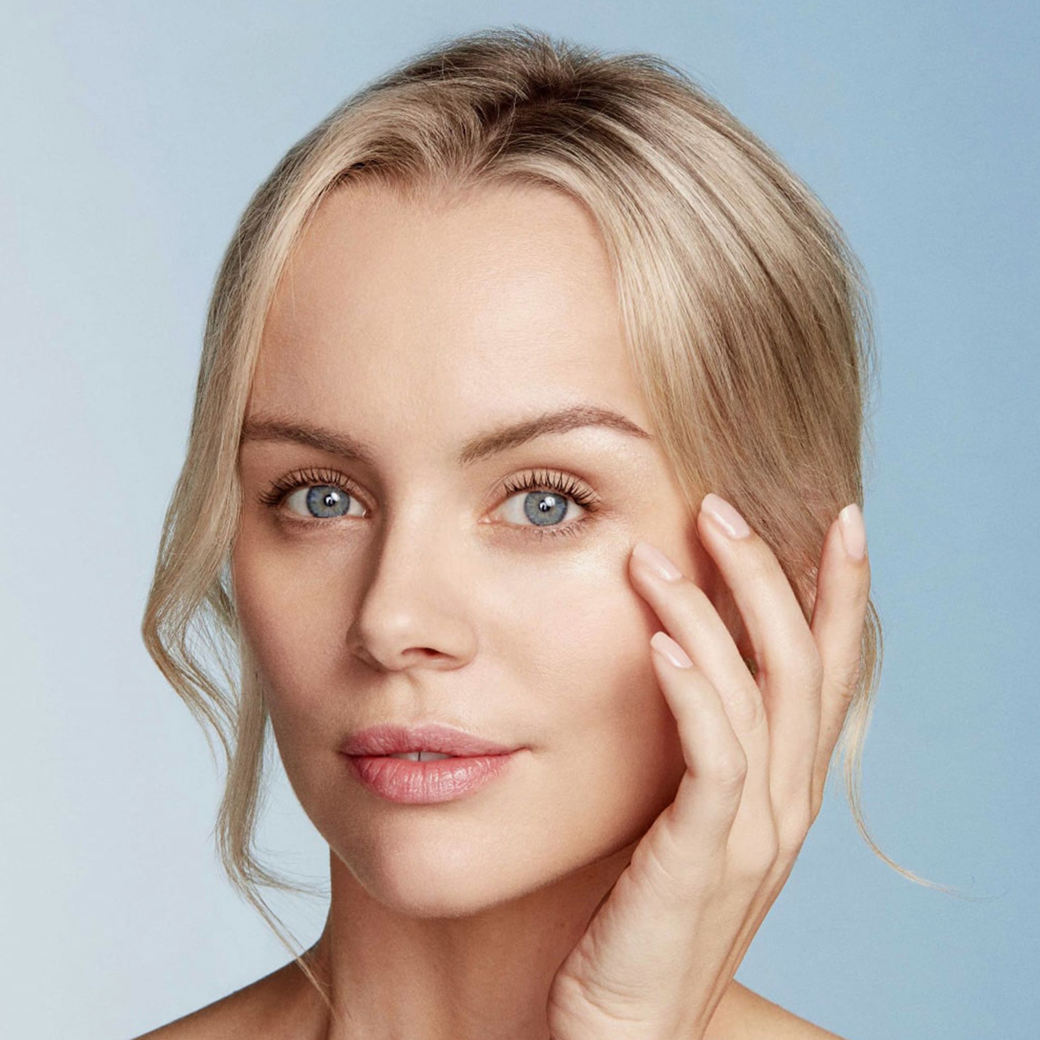 Frau mit blonden Haaren berührt sanft die Haut unter dem Auge. Blauer Hintergrund. Keine Produktbezeichnung sichtbar.