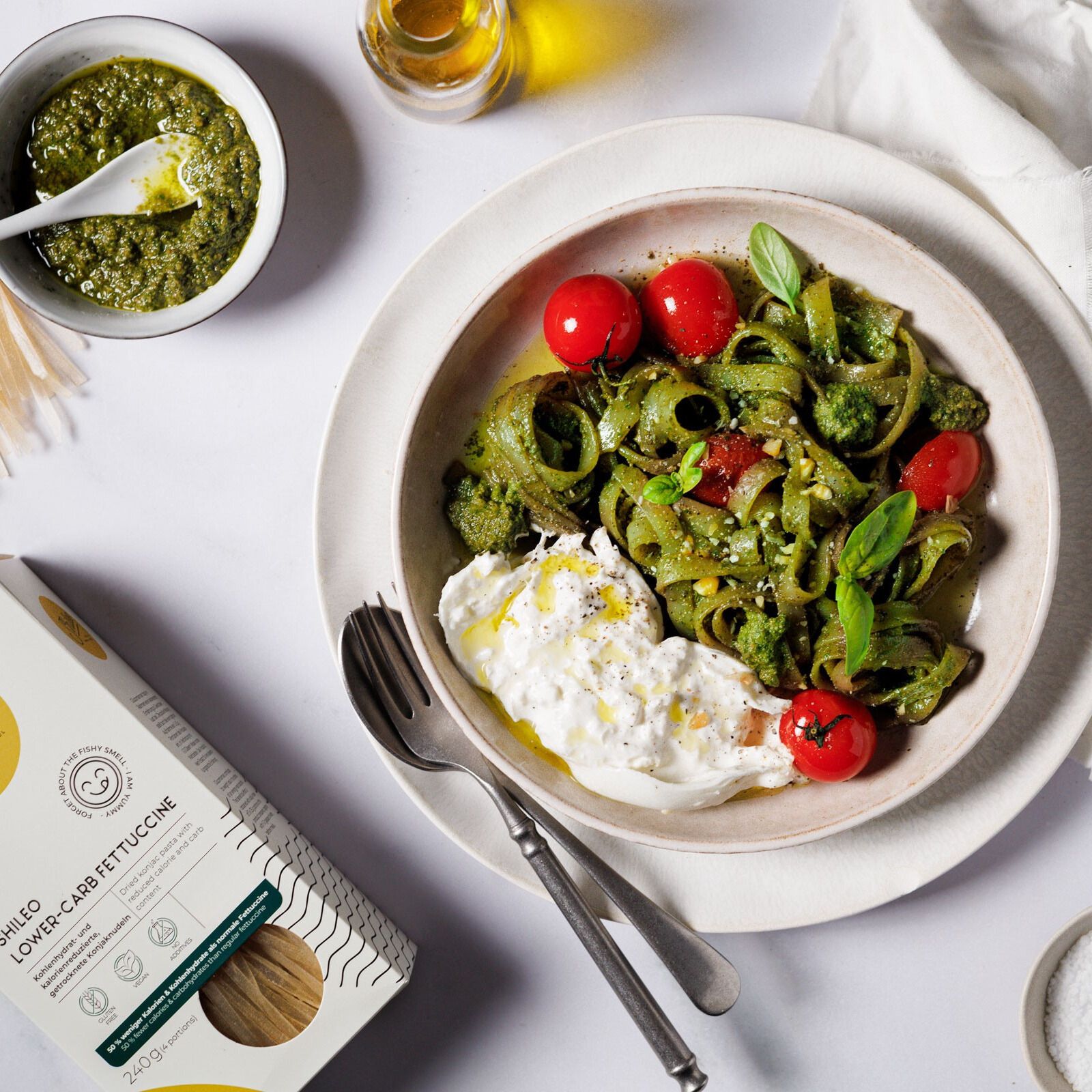 Teller mit grünen Nudeln, Tomaten und Käse. SHILEO Lower-Carb Fettuccine Packung links. Pesto und Öl.