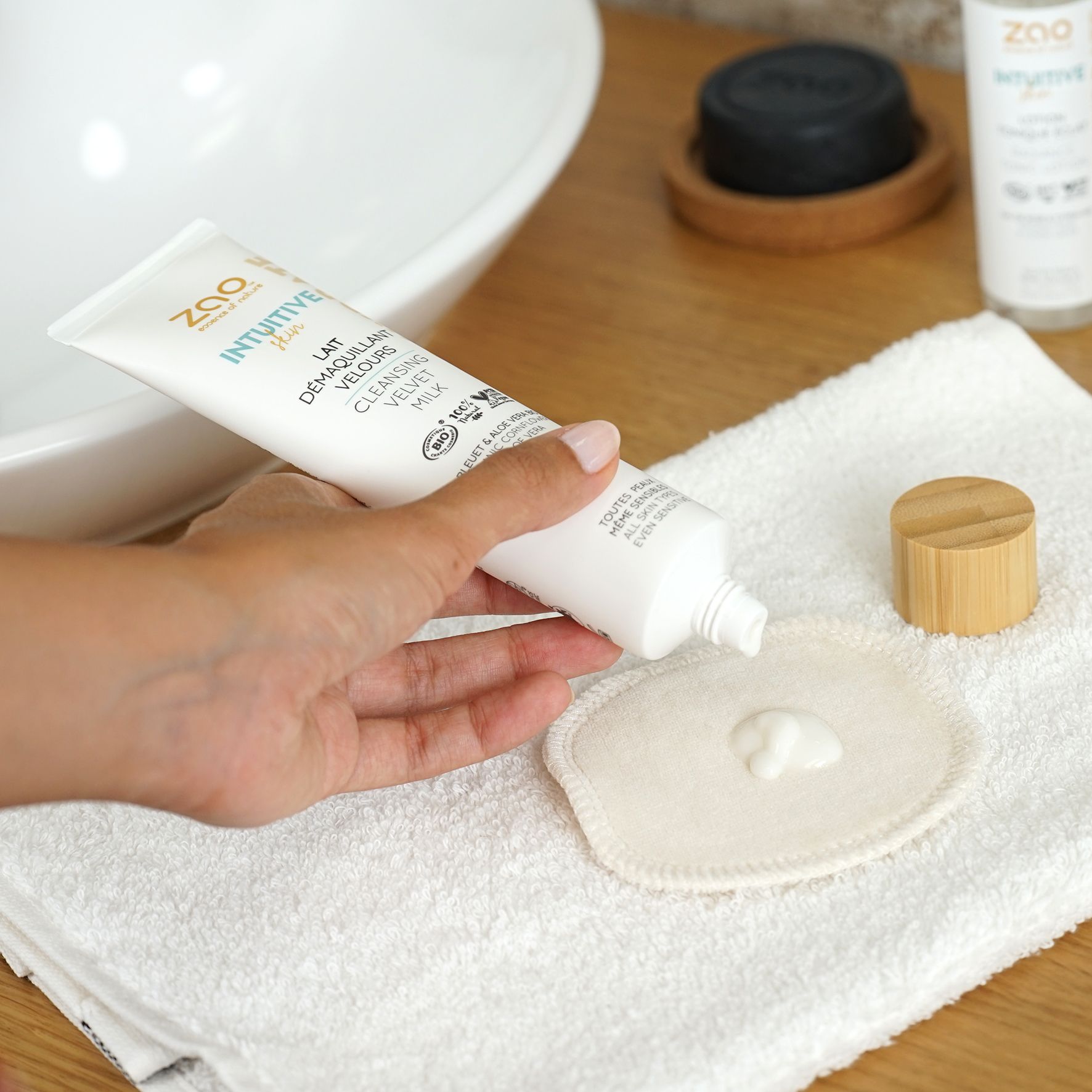 Weiße Tube mit Produkt, Text und Logo. Produkt wird auf Wattepad gedrückt. Auf Handtuch und neben Waschbecken.