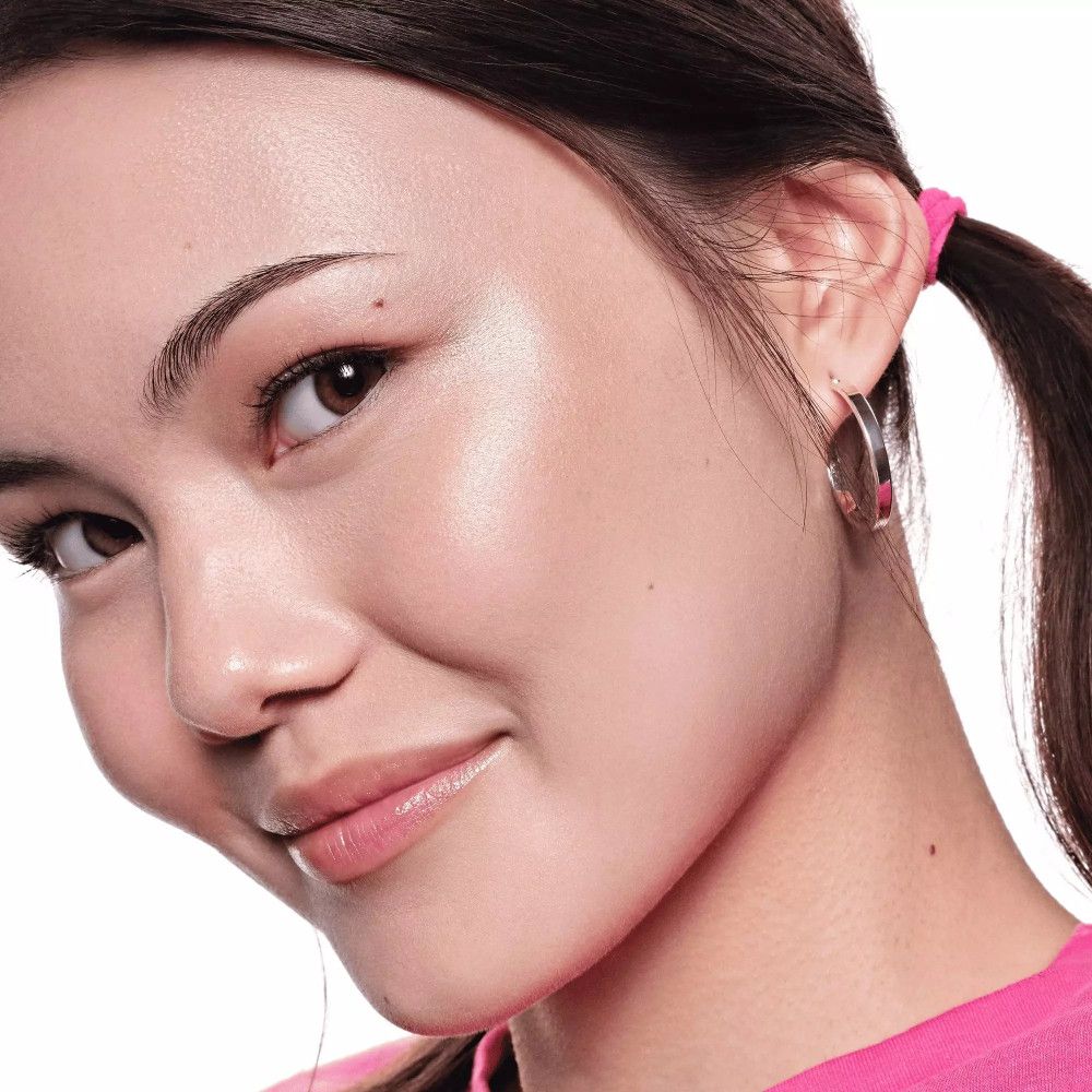 Nahaufnahme einer Frau mit gehighlighteten Wangenknochen. Sie trägt ein rosa Oberteil. Ihr Haar ist zu einem Pferdeschwanz gebunden.