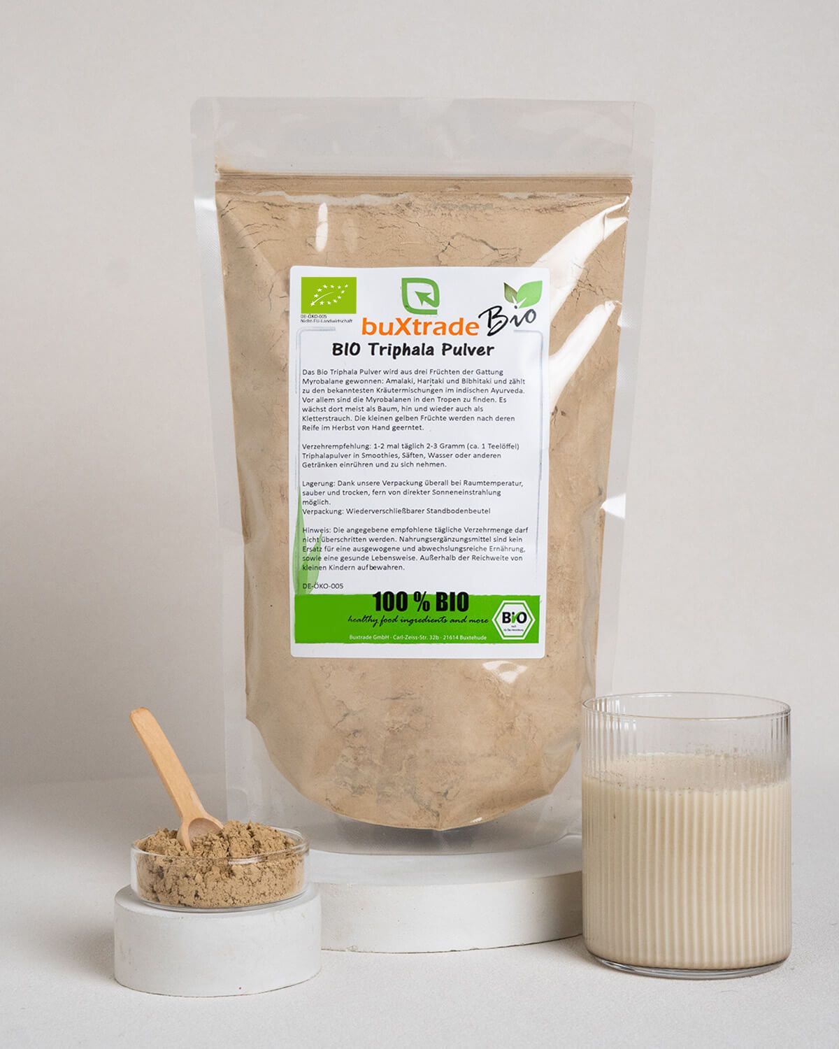 Bio Triphala Pulver in einem Standbodenbeutel mit Etikett. Daneben eine kleine Schale mit Pulver und ein Glas Milch.