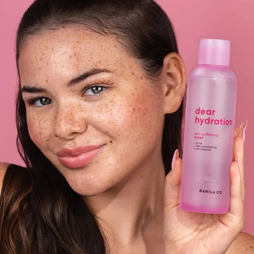 Eine Frau hält eine durchsichtige rosa Flasche mit der Aufschrift „dear hydration skin softening toner“ und „BANILA CO“.