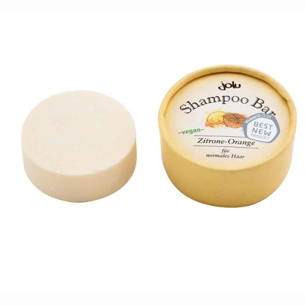 Runder, heller Shampoo Bar neben einer runden Pappverpackung. Auf der Verpackung steht "Shampoo Bar Zitrone-Orange" und "vegan".