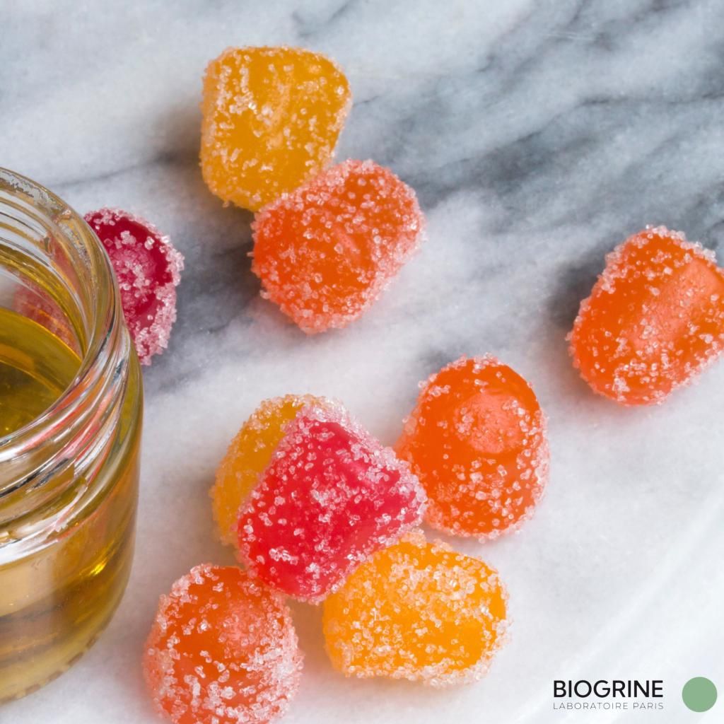 CBD Gummies auf Marmor. Einige liegen neben einem Glas. BIOGRINE Logo unten rechts.