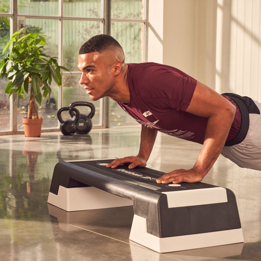 Schwarzer Reebok Step mit weißen Füßen. Mann macht Liegestütze darauf. Im Hintergrund: Fenster, Pflanze, Hanteln.