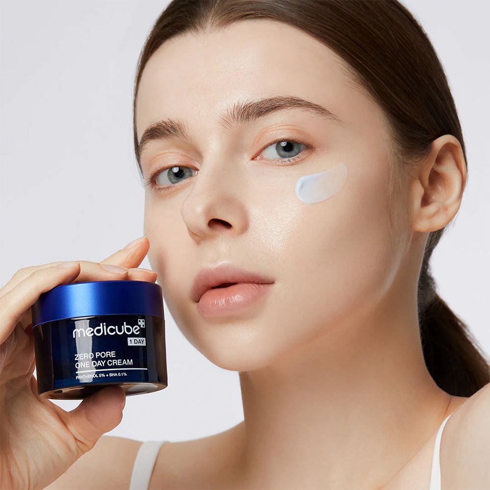 Frau hält Medicube Zero Pore One Day Cream Tiegel. Ein Tupfer Creme auf ihrer Wange.