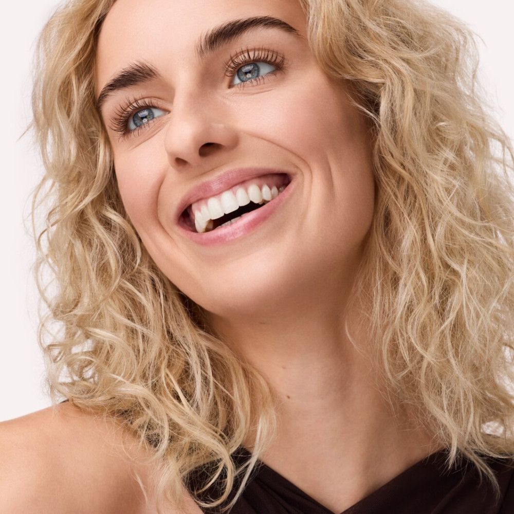 Porträt einer lächelnden Frau mit blonden Locken und blauen Augen. Sie trägt dunkle Kleidung.