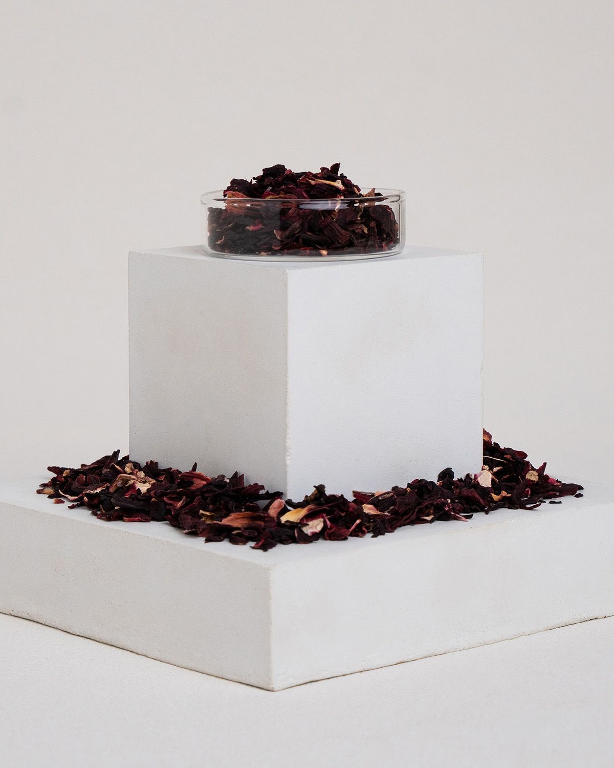 Kleine Schale mit Hibiskusblüten auf weißem Podest. Blüten verstreut. Hintergrund weiß.